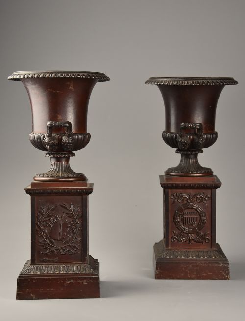fig 2. A pair of dark brown patinated spelter campana urns featuring gadrooned bases, scroll handles, and square stepped plinths with relief decoration.