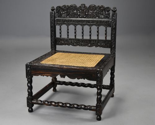 fig 1. A 17th-century dark carved ebony low chair featuring floral relief carving on the frame, a hand-woven cane seat, and spiralled barley-twist legs.