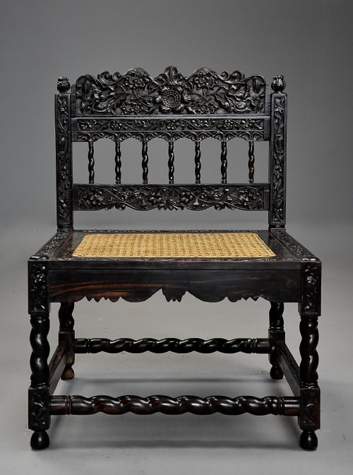 fig 2. A 17th-century dark carved ebony low chair featuring floral relief carving on the frame, a hand-woven cane seat, and spiralled barley-twist legs.