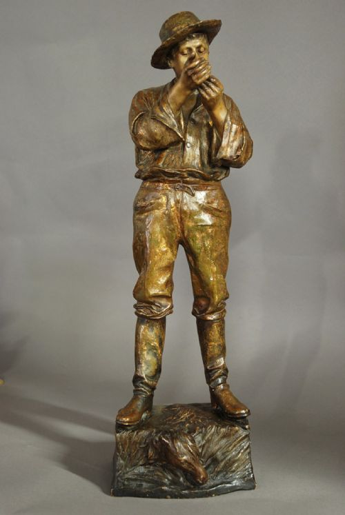 fig 1. A bronzed terracotta figure of a standing man wearing a hat and lighting a pipe on a naturalistic base.