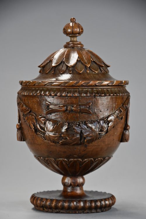 fig 1. A late eighteenth-century carved walnut lidded urn featuring floral garlands, tassel motifs, a leaf-carved dome lid with finial, resting on a turned circular base.