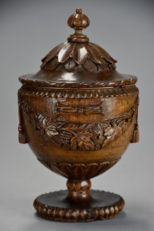 fig 2. A late eighteenth-century carved walnut lidded urn featuring floral garlands, tassel motifs, a leaf-carved dome lid with finial, resting on a turned circular base.