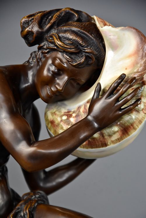fig 4. A patinated dark bronze sculpture depicting a youth kneeling and holding a large seashell to his ear on an octagonal base.
