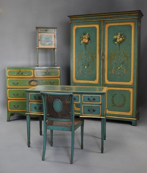 fig 1. An early twentieth-century hand-painted green and gold bedroom suite featuring a wardrobe, chest of drawers with mirror, dressing desk, side tables, and caned chairs decorated with floral urn motifs.
