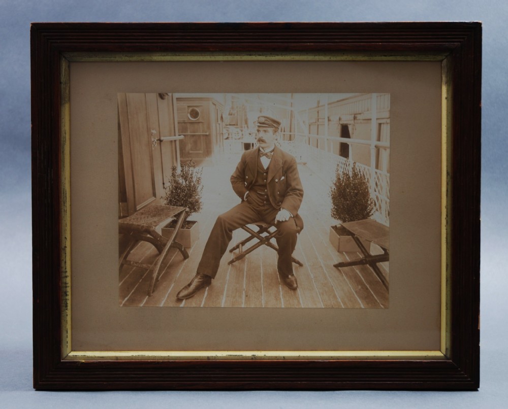 An early twentieth century framed sepia photograph depicting a ship captain seated on the wooden deck of a vessel.