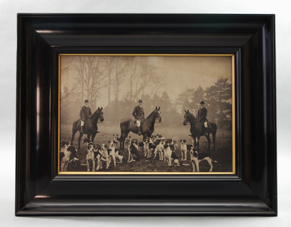 A framed Victorian monochrome photograph depicting three huntsmen mounted on horses surrounded by a pack of foxhounds.