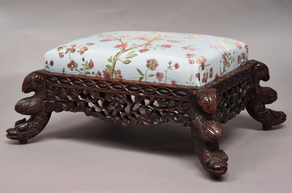 A late 19th-century Anglo-Indian dark hardwood footstool with intricately carved floral aprons, zoomorphic legs, and a light blue floral upholstered seat.