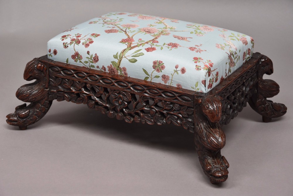 A late 19th-century Anglo-Indian dark hardwood footstool with intricately carved floral aprons, zoomorphic legs, and a light blue floral upholstered seat. View II