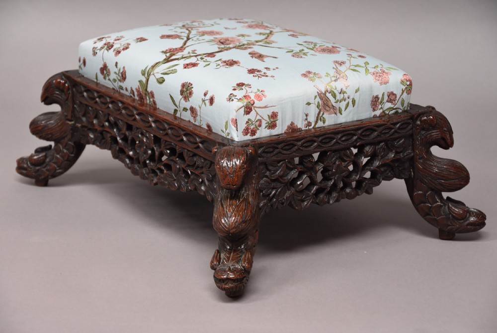 A late 19th-century Anglo-Indian dark hardwood footstool with intricately carved floral aprons, zoomorphic legs, and a light blue floral upholstered seat. View V