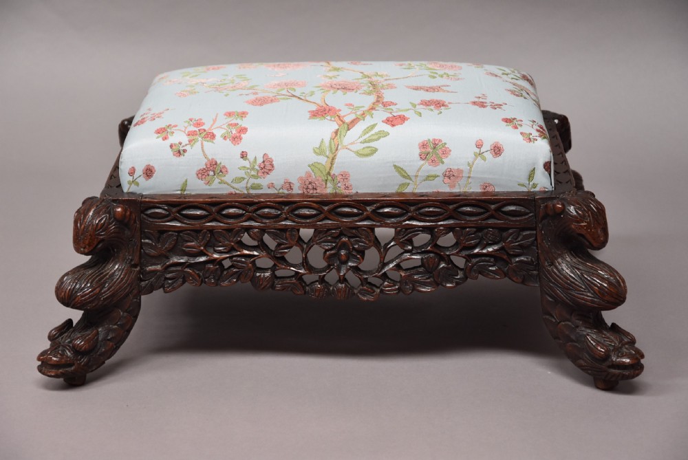 A late 19th-century Anglo-Indian dark hardwood footstool with intricately carved floral aprons, zoomorphic legs, and a light blue floral upholstered seat. View VI