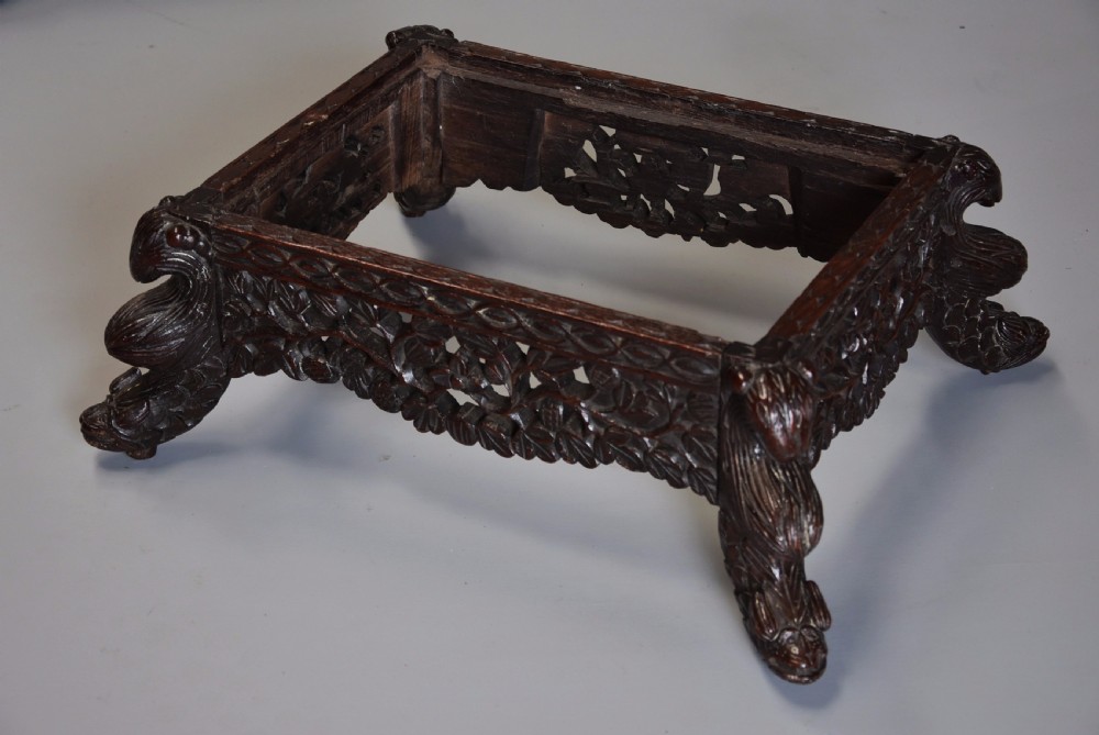 A late 19th-century Anglo-Indian dark hardwood footstool with intricately carved floral aprons, zoomorphic legs, and a light blue floral upholstered seat. View X