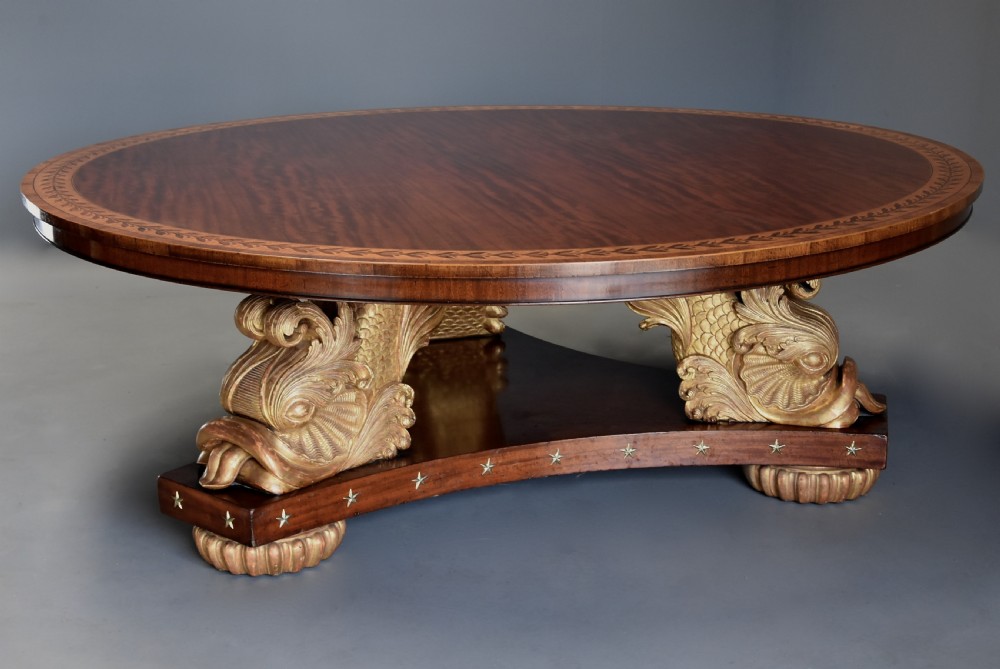 A large circular Regency style mahogany centre table featuring a crossbanded marquetry top and a tri-form base supported by three carved gilded dolphin figures with brass star details.