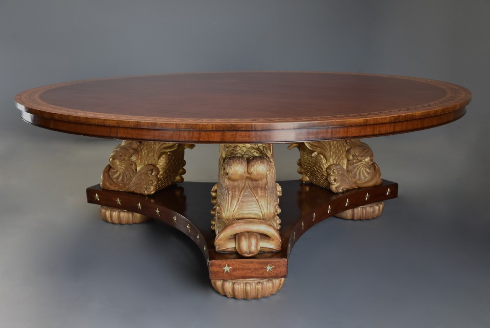 A large circular Regency style mahogany centre table featuring a crossbanded marquetry top and a tri-form base supported by three carved gilded dolphin figures with brass star details. View II