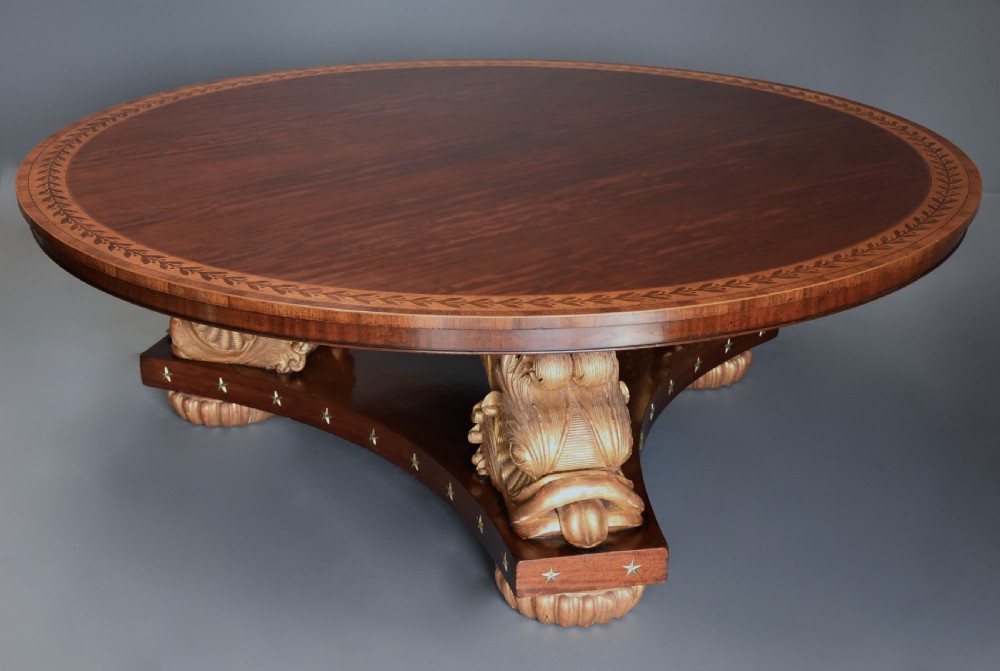 A large circular Regency style mahogany centre table featuring a crossbanded marquetry top and a tri-form base supported by three carved gilded dolphin figures with brass star details. View III