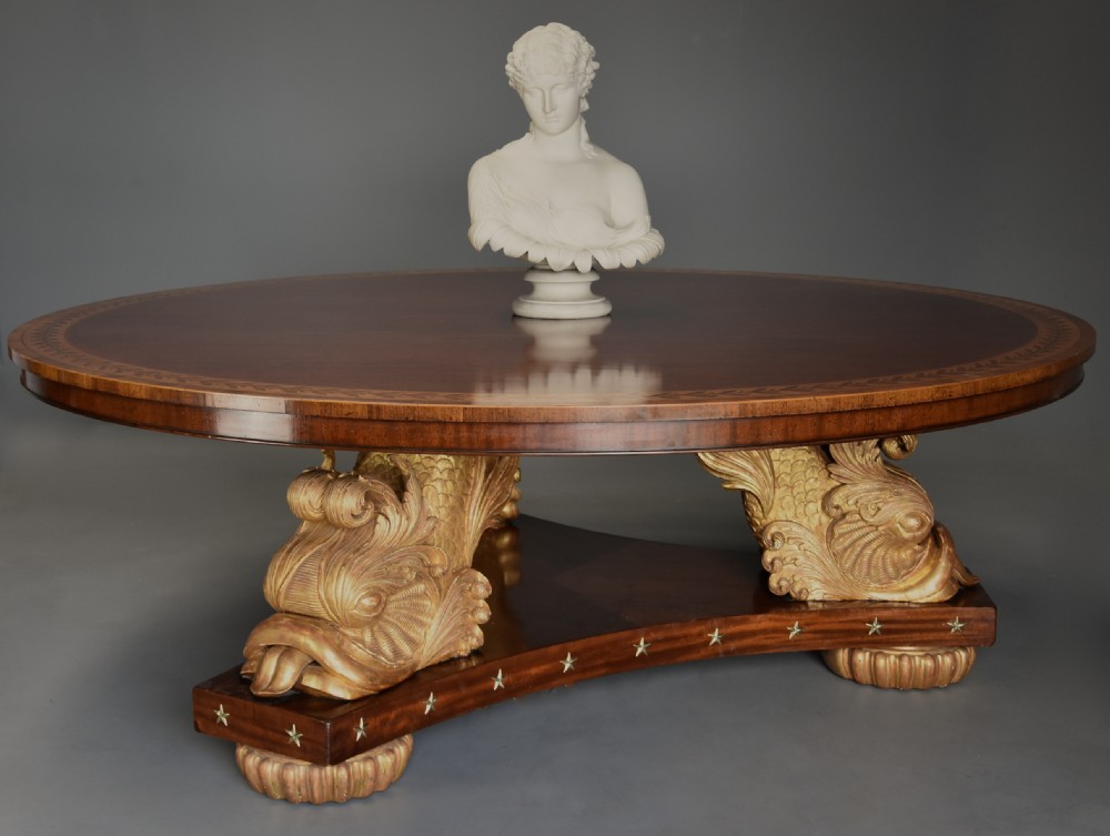 A large circular Regency style mahogany centre table featuring a crossbanded marquetry top and a tri-form base supported by three carved gilded dolphin figures with brass star details. View V