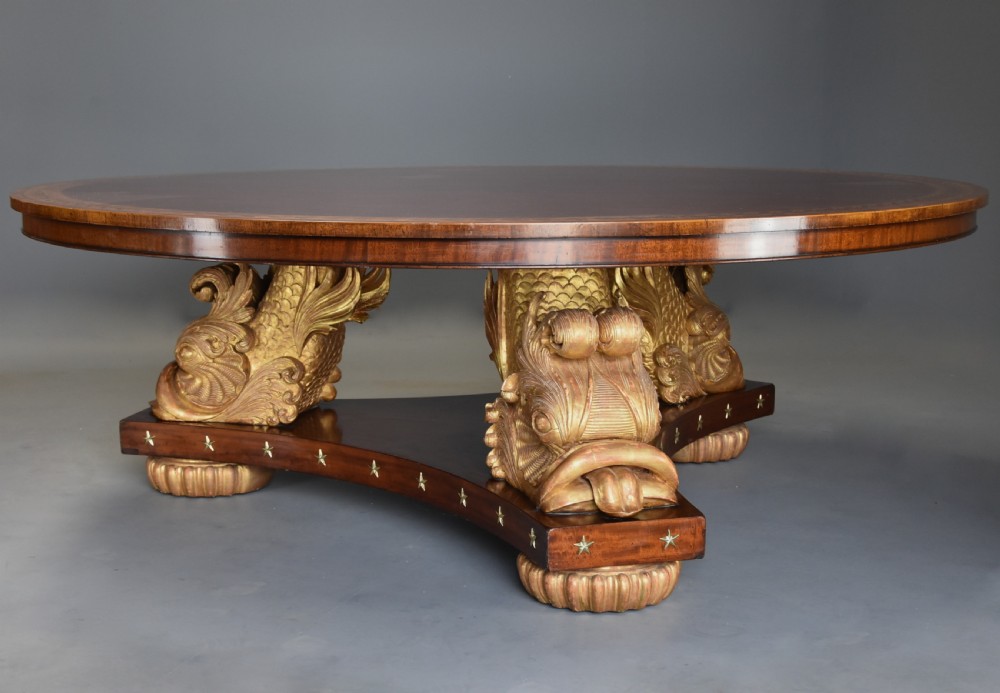 A large circular Regency style mahogany centre table featuring a crossbanded marquetry top and a tri-form base supported by three carved gilded dolphin figures with brass star details. View VI