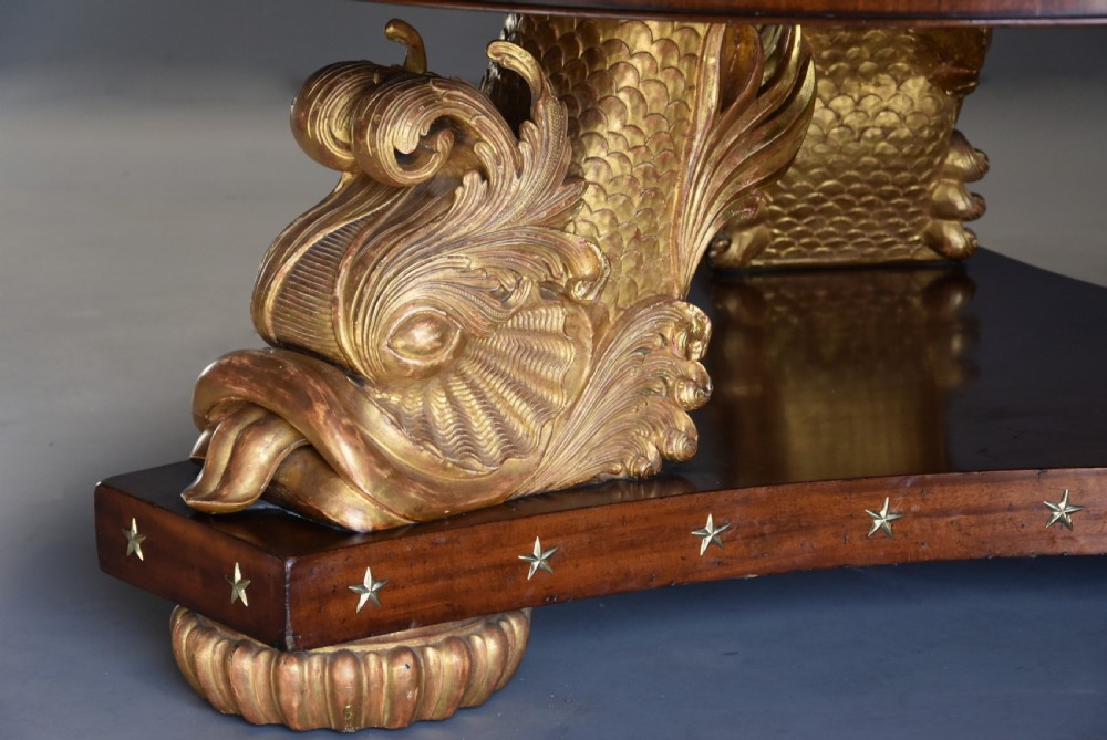 A large circular Regency style mahogany centre table featuring a crossbanded marquetry top and a tri-form base supported by three carved gilded dolphin figures with brass star details. View VIII