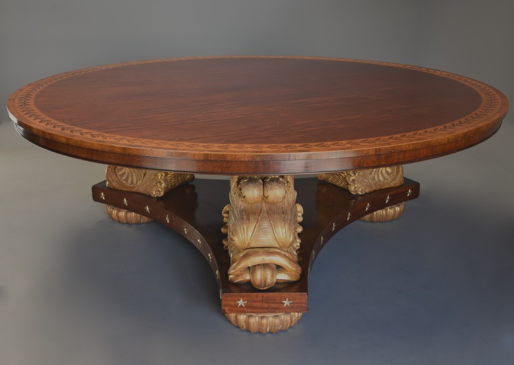 A large circular Regency style mahogany centre table featuring a crossbanded marquetry top and a tri-form base supported by three carved gilded dolphin figures with brass star details. View IX