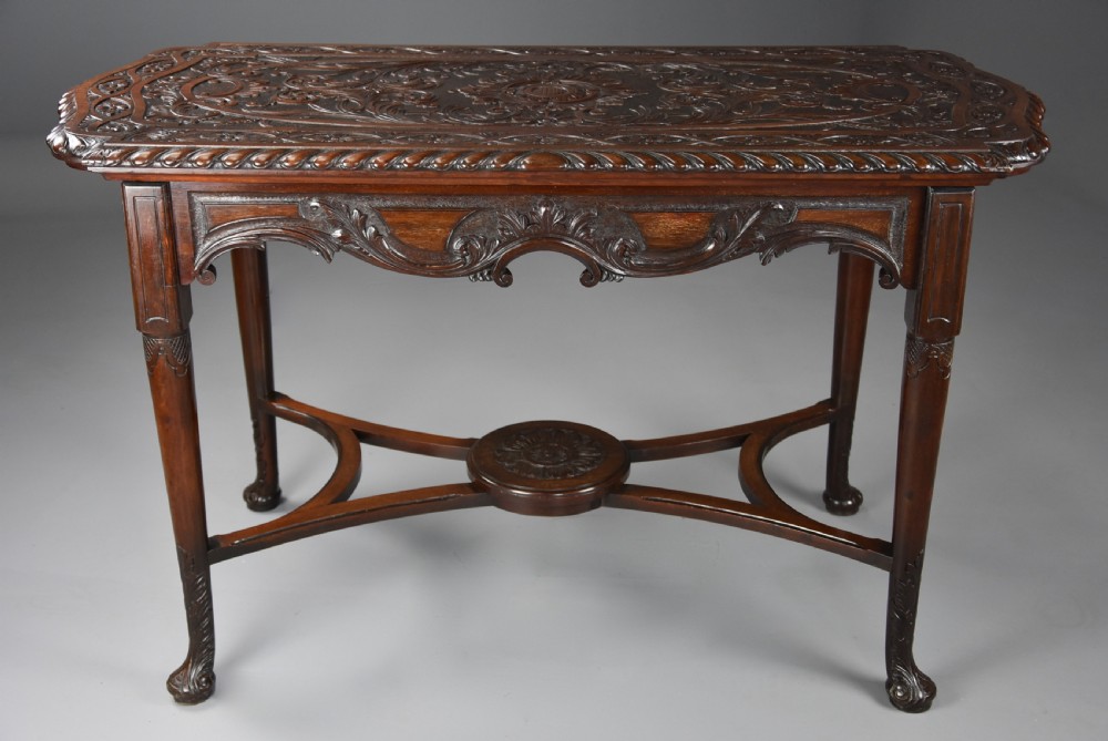 An antique dark mahogany centre table featuring intricate relief carvings on the top, a shaped carved apron, and an X-frame stretcher with a central roundel.