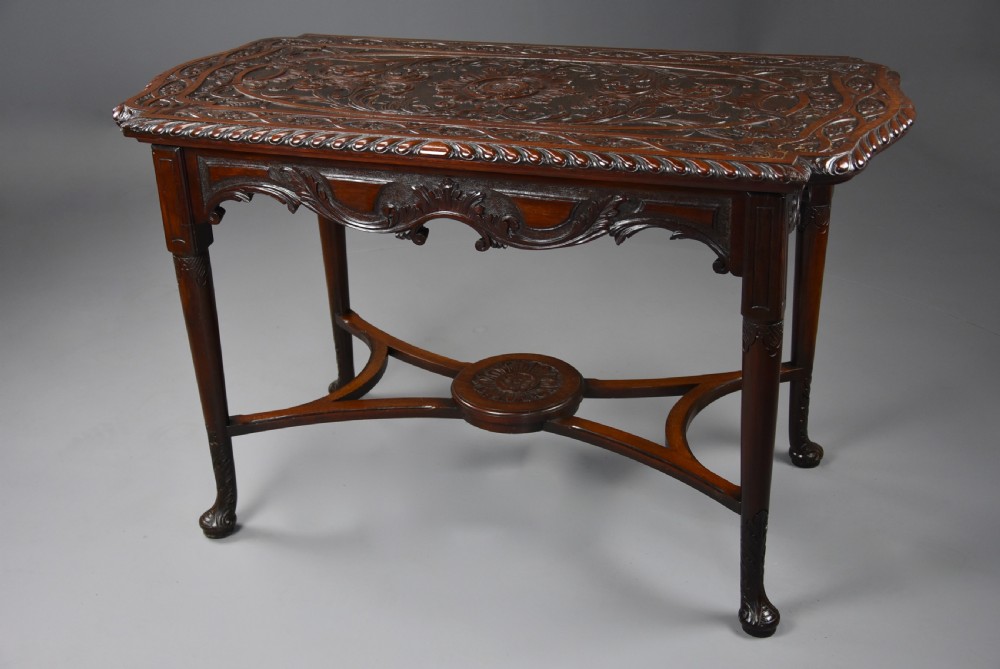 An antique dark mahogany centre table featuring intricate relief carvings on the top, a shaped carved apron, and an X-frame stretcher with a central roundel. View IV