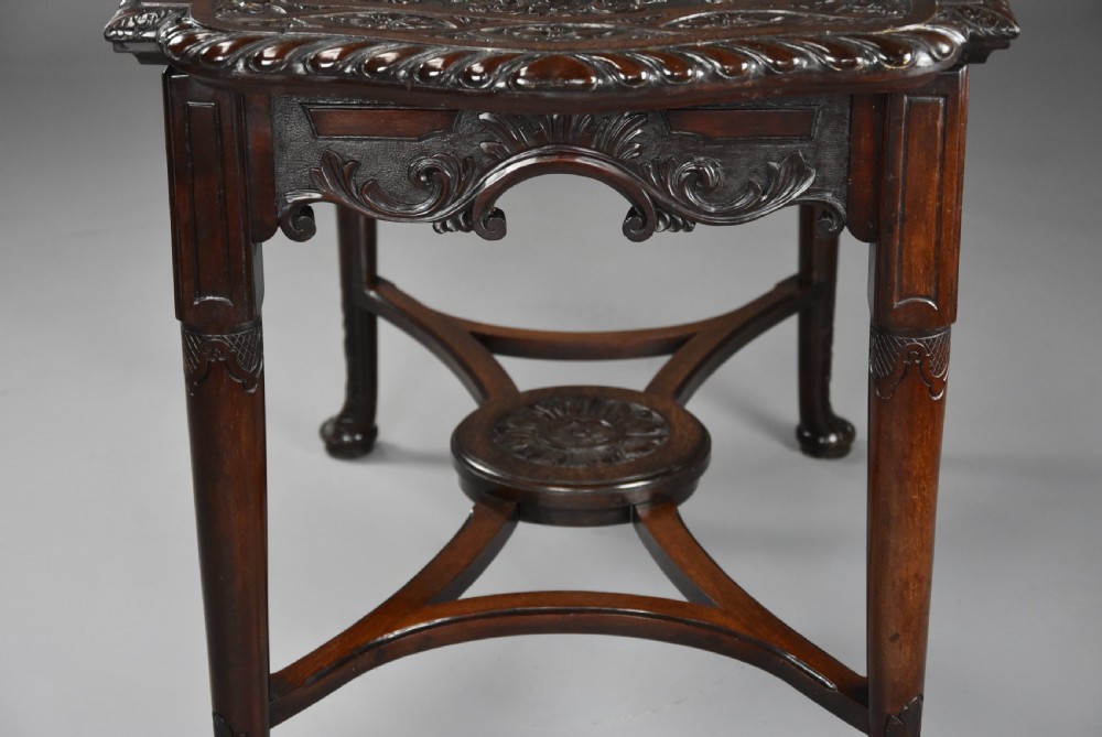 An antique dark mahogany centre table featuring intricate relief carvings on the top, a shaped carved apron, and an X-frame stretcher with a central roundel. View VI