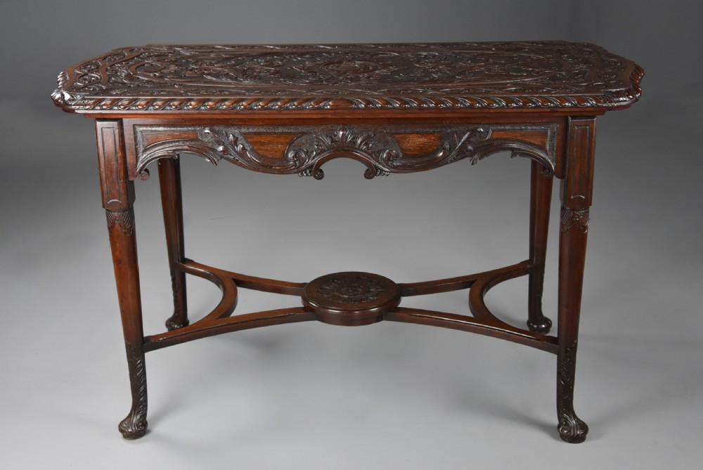 An antique dark mahogany centre table featuring intricate relief carvings on the top, a shaped carved apron, and an X-frame stretcher with a central roundel. View VIII