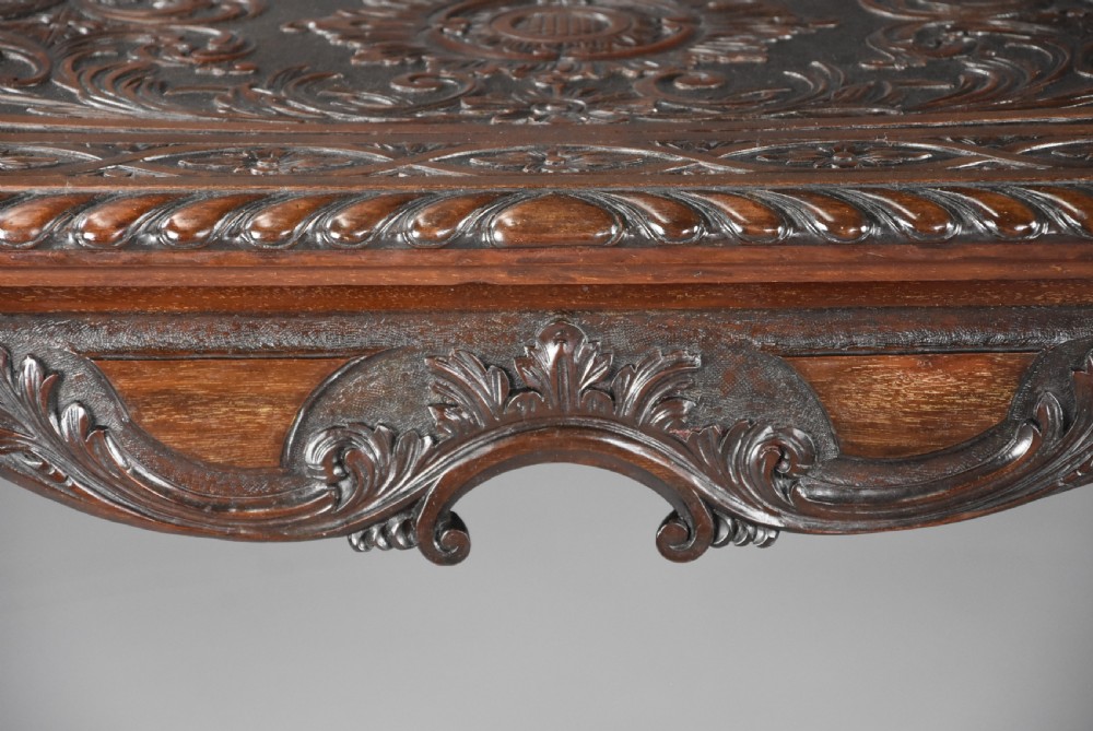 An antique dark mahogany centre table featuring intricate relief carvings on the top, a shaped carved apron, and an X-frame stretcher with a central roundel. View IX