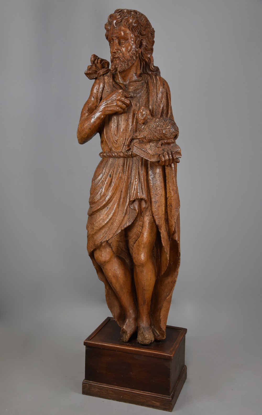 A 16th-century carved limewood figure of St. John the Baptist holding a lamb, mounted on a dark wooden base.