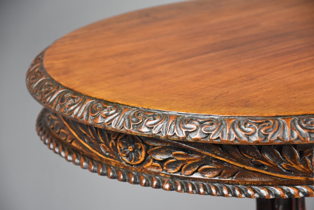 A mid 19th-century Anglo-Indian circular tilt-top occasional table crafted from dark carved padouk wood with a foliate apron, turned pedestal, and tripartite base. View VIII