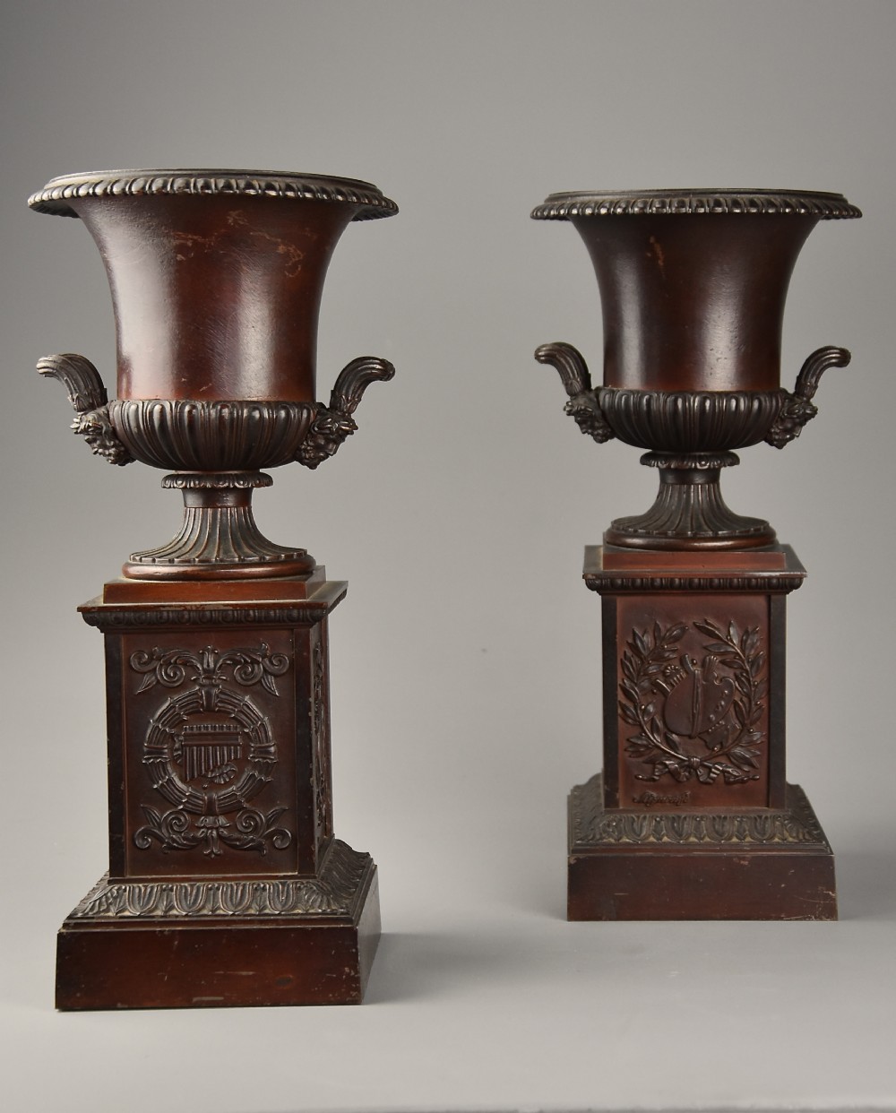 A pair of dark brown patinated spelter campana urns featuring gadrooned bases, scroll handles, and square stepped plinths with relief decoration.
