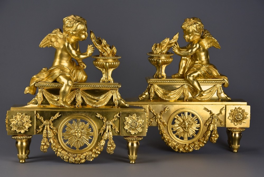 A pair of late nineteenth-century gilded bronze fire dogs featuring seated winged cherubs warming their hands by flaming urns above solar masks and drapery.