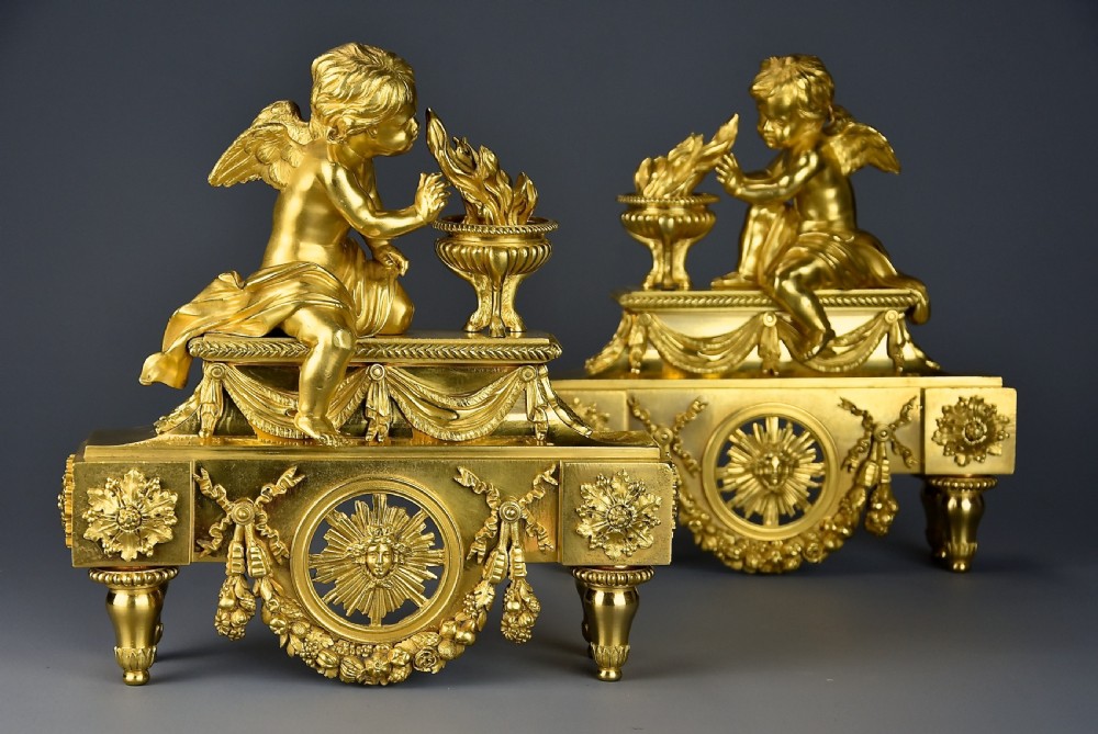 A pair of late nineteenth-century gilded bronze fire dogs featuring seated winged cherubs warming their hands by flaming urns above solar masks and drapery. View II