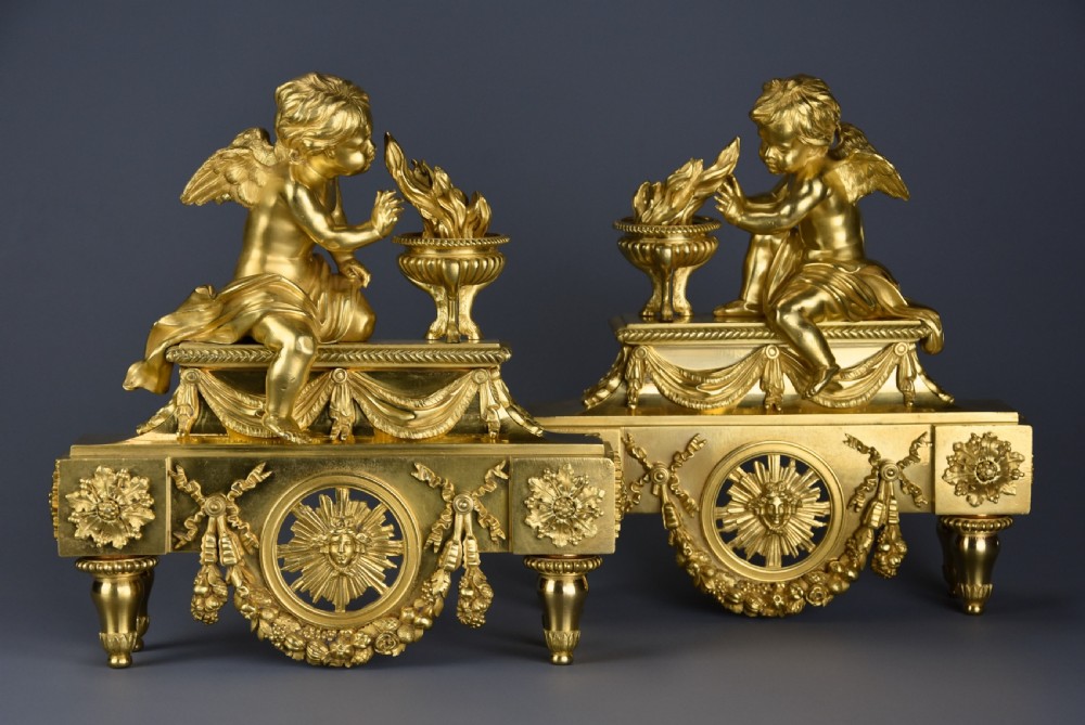 A pair of late nineteenth-century gilded bronze fire dogs featuring seated winged cherubs warming their hands by flaming urns above solar masks and drapery. View III
