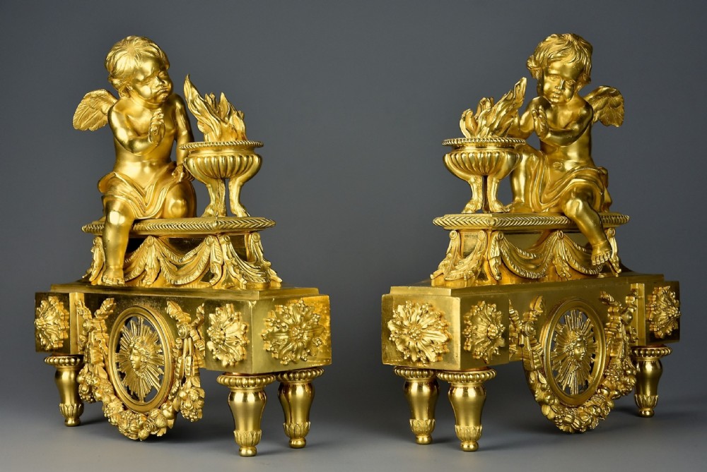 A pair of late nineteenth-century gilded bronze fire dogs featuring seated winged cherubs warming their hands by flaming urns above solar masks and drapery. View V