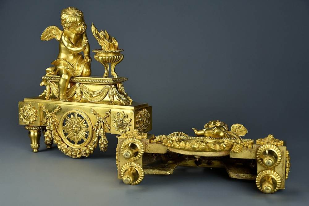 A pair of late nineteenth-century gilded bronze fire dogs featuring seated winged cherubs warming their hands by flaming urns above solar masks and drapery. View XII
