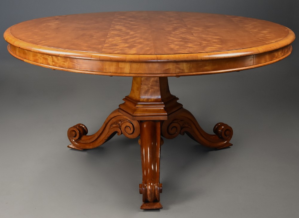 A circular mid-19th-century tilt-top breakfast table made from figured satin birch timber with a carved central pedestal base and scroll feet.