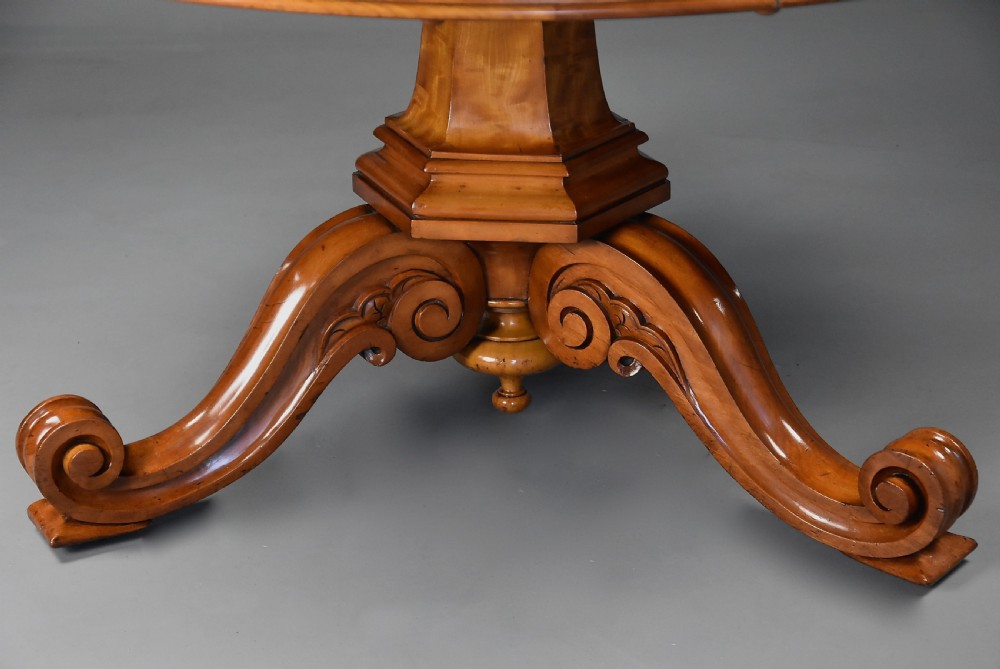A circular mid-19th-century tilt-top breakfast table made from figured satin birch timber with a carved central pedestal base and scroll feet. View IX