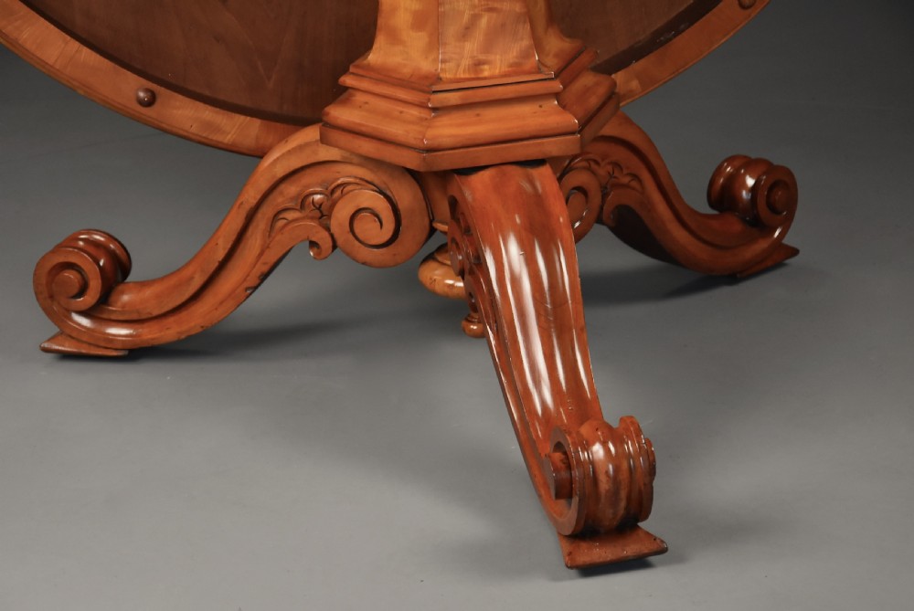 A circular mid-19th-century tilt-top breakfast table made from figured satin birch timber with a carved central pedestal base and scroll feet. View X