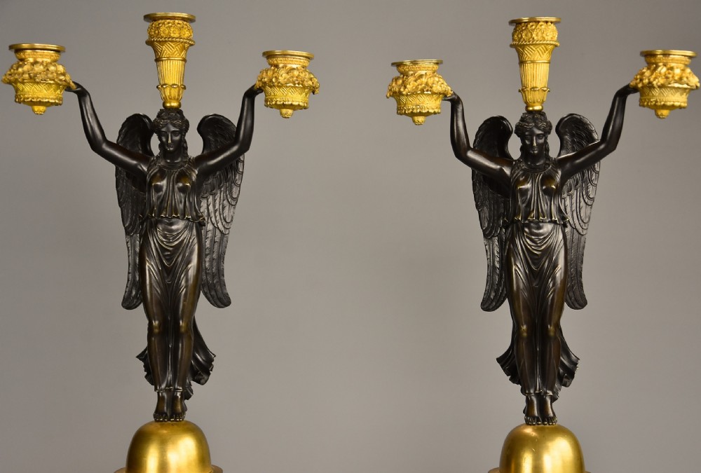 A pair of early nineteenth-century French Empire patinated and gilt bronze candelabra featuring winged classical figures holding candle nozzles aloft on decorated cylindrical pedestals. View IV