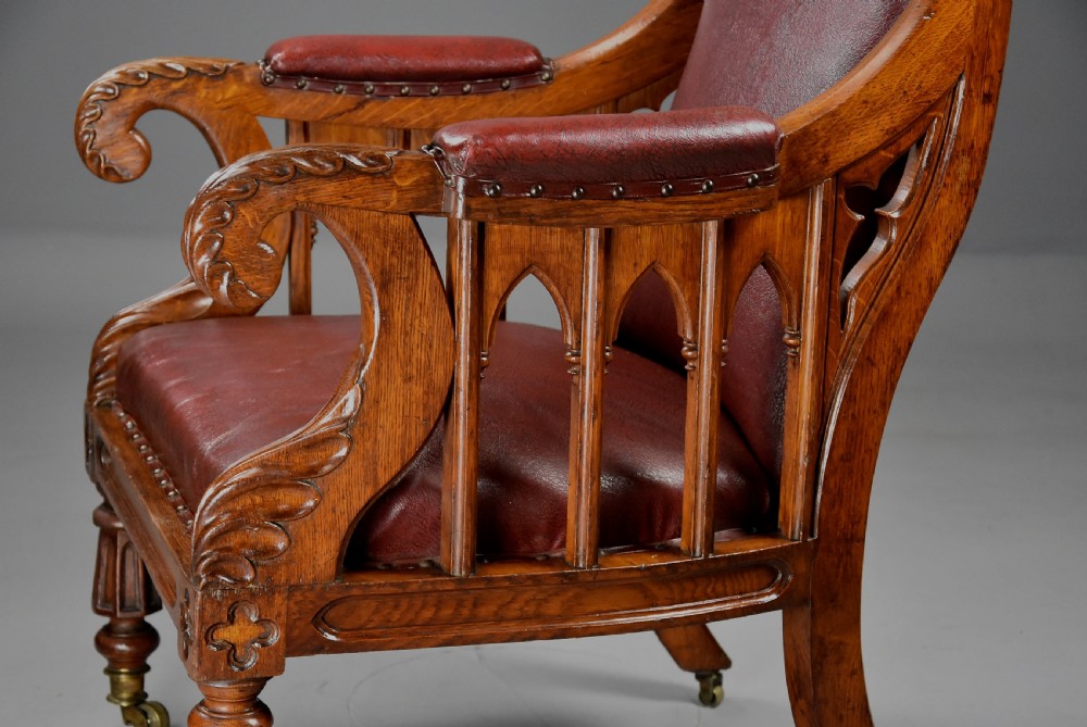 A 19th-century Gothic Revival carved oak armchair upholstered in deep red leather with tracery sides and turned front legs on brass castors. View VII