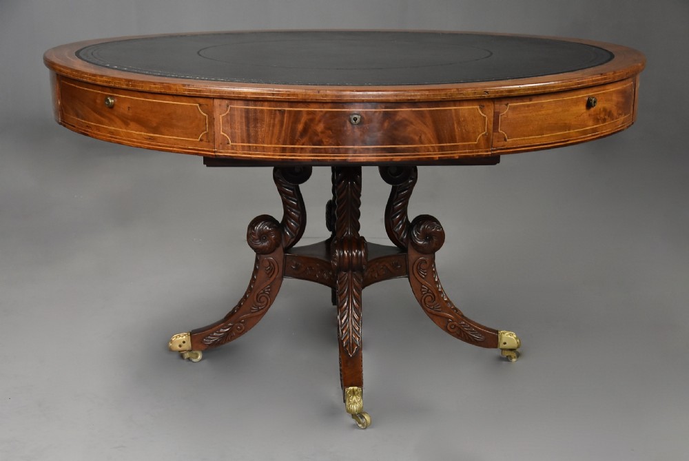 An early 19th-century circular mahogany Regency drum table featuring a dark leather inset top, frieze drawers with line inlay, and a carved central pedestal supported by four scroll legs on brass castors. View IV