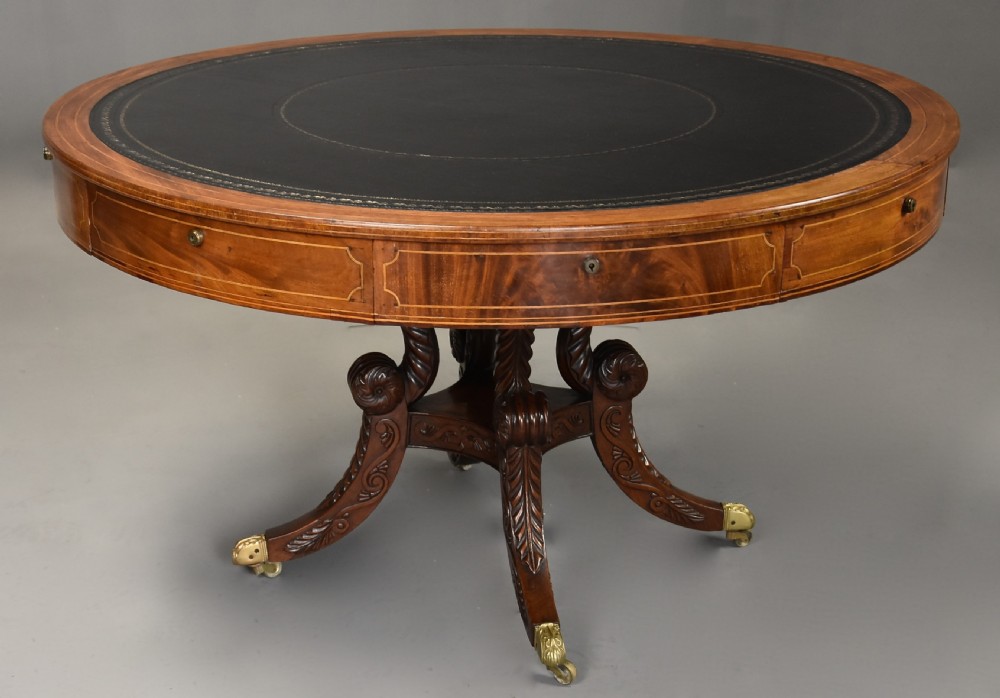 An early 19th-century circular mahogany Regency drum table featuring a dark leather inset top, frieze drawers with line inlay, and a carved central pedestal supported by four scroll legs on brass castors. View V
