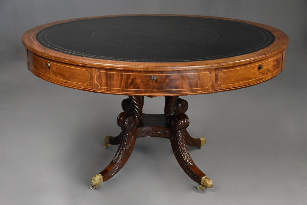 An early 19th-century circular mahogany Regency drum table featuring a dark leather inset top, frieze drawers with line inlay, and a carved central pedestal supported by four scroll legs on brass castors. View VI