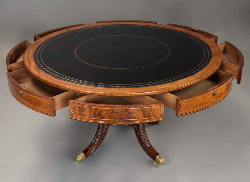 An early 19th-century circular mahogany Regency drum table featuring a dark leather inset top, frieze drawers with line inlay, and a carved central pedestal supported by four scroll legs on brass castors. View X
