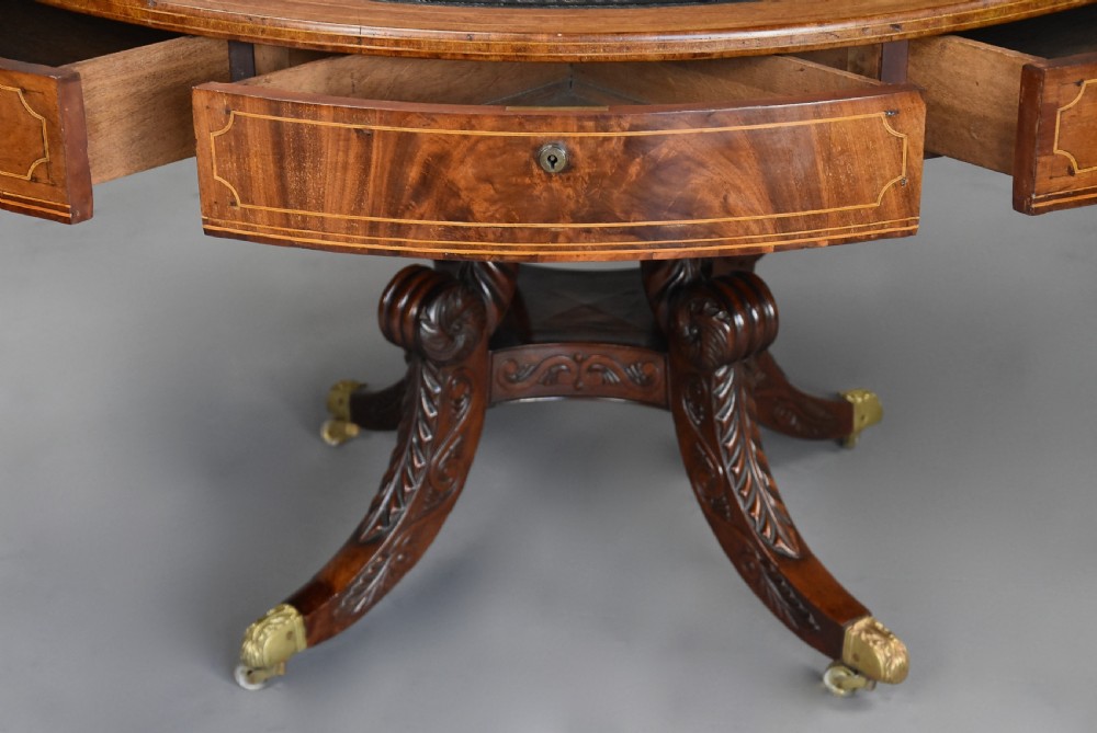 An early 19th-century circular mahogany Regency drum table featuring a dark leather inset top, frieze drawers with line inlay, and a carved central pedestal supported by four scroll legs on brass castors. View XI