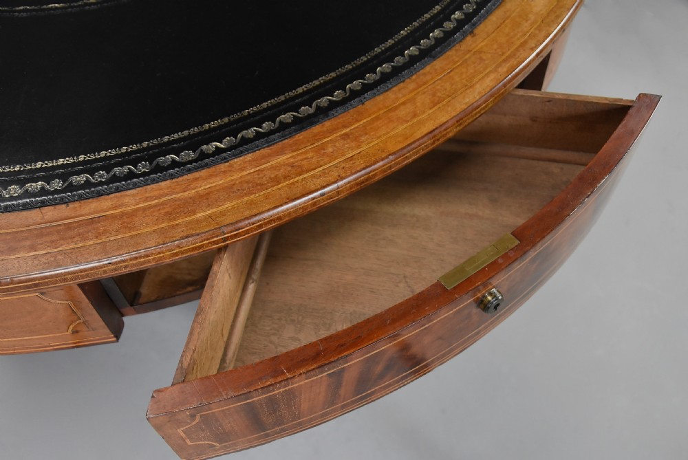 An early 19th-century circular mahogany Regency drum table featuring a dark leather inset top, frieze drawers with line inlay, and a carved central pedestal supported by four scroll legs on brass castors. View XII