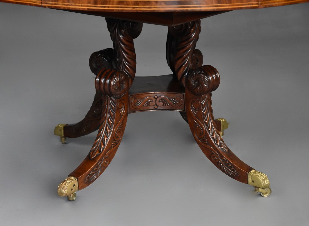An early 19th-century circular mahogany Regency drum table featuring a dark leather inset top, frieze drawers with line inlay, and a carved central pedestal supported by four scroll legs on brass castors. View XIV