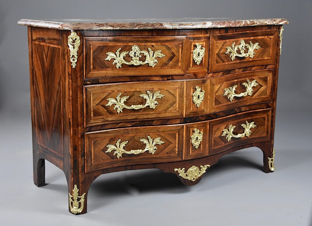 An 18th-century French Regence serpentine commode crafted in rosewood parquetry with gilded ormolu drawer pulls and a contoured marble top.