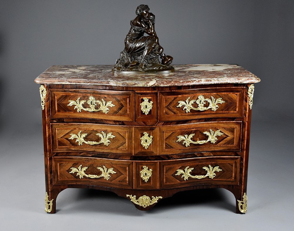 An 18th-century French Regence serpentine commode crafted in rosewood parquetry with gilded ormolu drawer pulls and a contoured marble top. View V