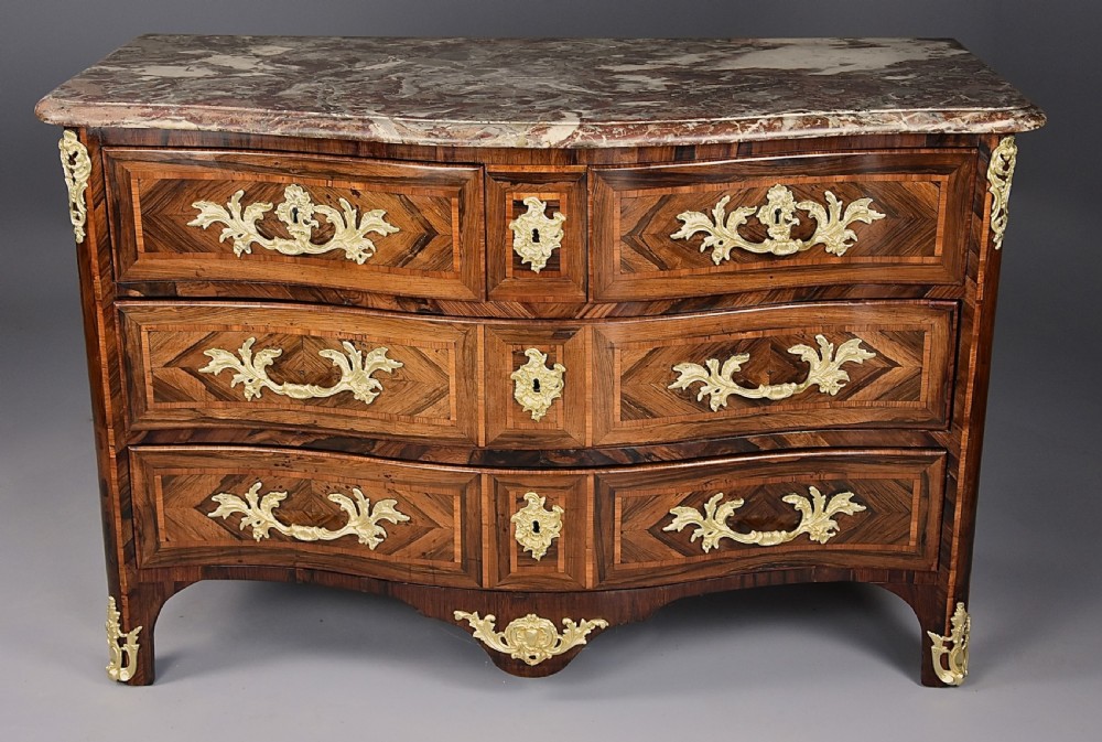 An 18th-century French Regence serpentine commode crafted in rosewood parquetry with gilded ormolu drawer pulls and a contoured marble top. View VI